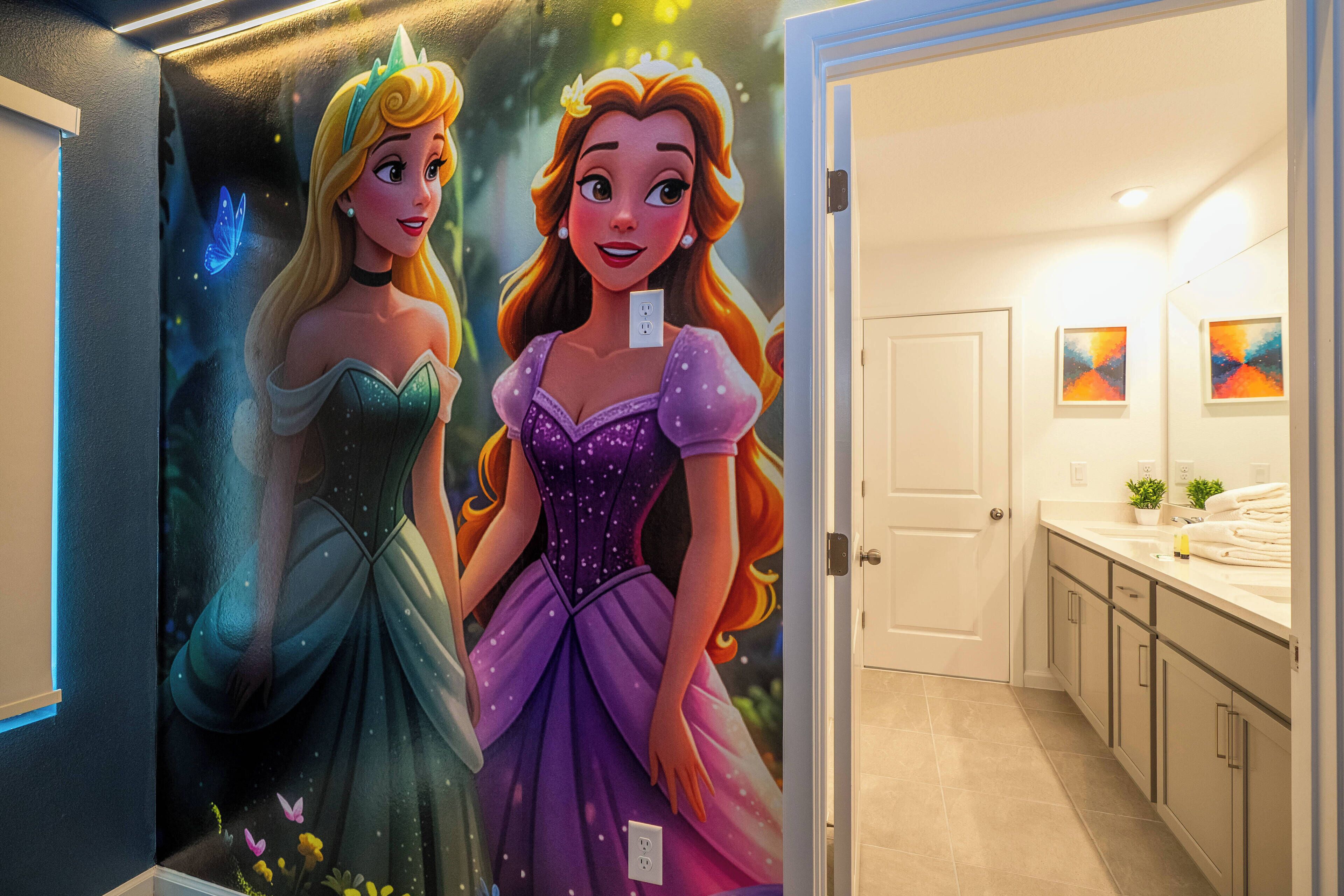Une salle de bain avec une grande fresque représentant des princesses Disney, une porte blanche et un plan de toilette agrémenté d’une plante et de serviettes.