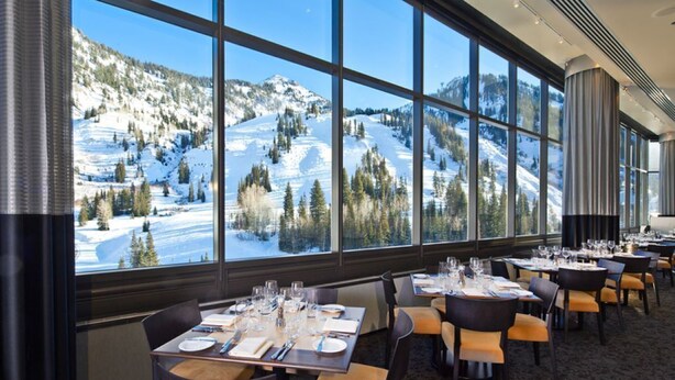 Ein Restaurant mit gedeckten Tischen und großen Fenstern, durch die sich eine verschneite Berglandschaft blickt.
