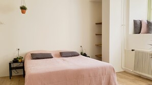 Een slaapkamer met een bed, twee nachtkastjes, een hangende plant, een radiator en een deur met planken.