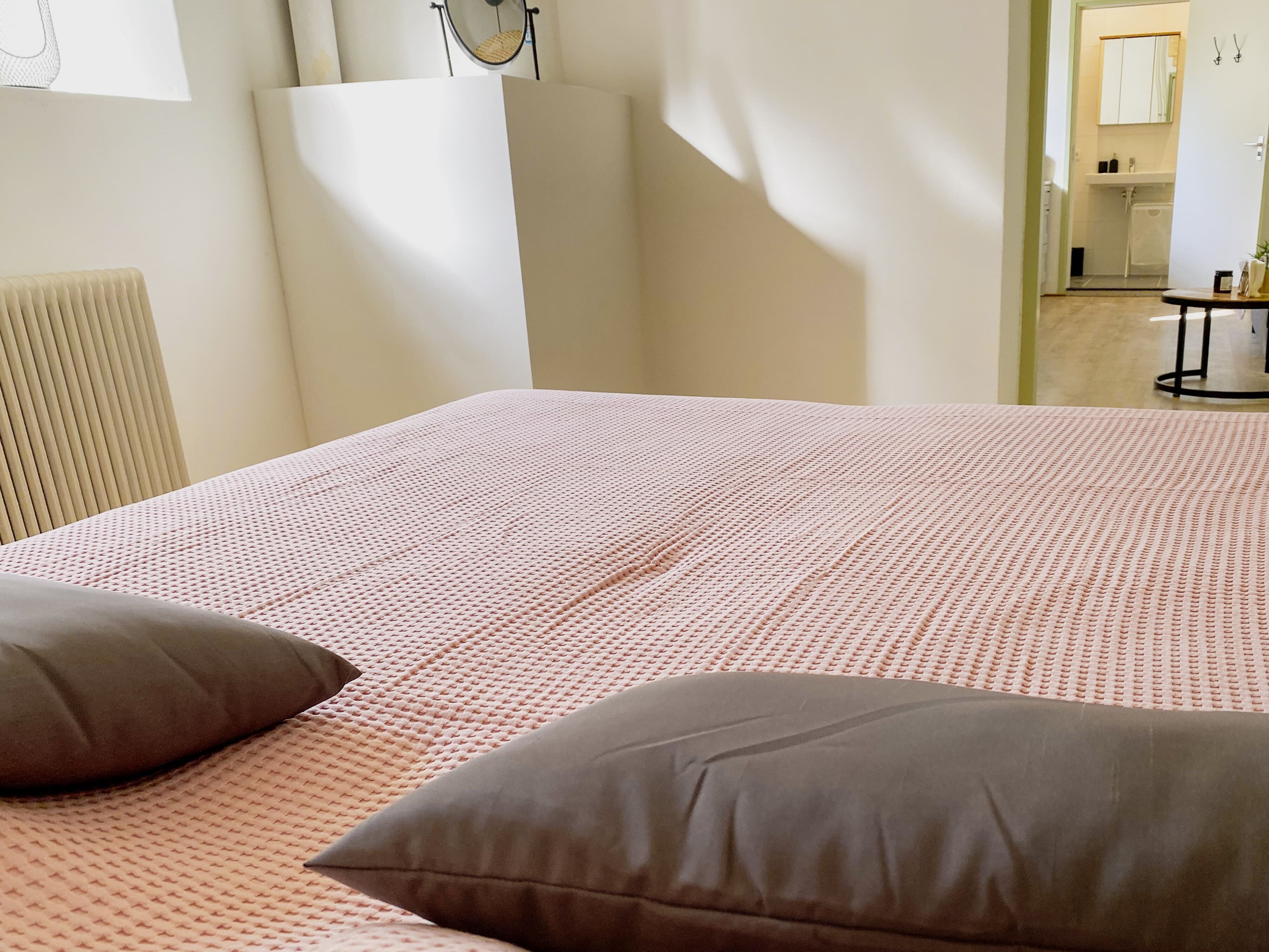 Een slaapkamer met een bed, kussens, een radiator en een badkamer die door een geopende deur zichtbaar is.