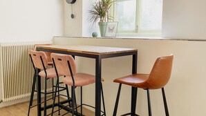 Een kamer met een houten tafel en vier stoelen, een radiator, een kamerplant en een raam met uitzicht.