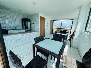 A modern kitchen with white tiles, a counter with a refrigerator, and a dining area with wicker chairs and a glass table. - Charming Studio Near the Airport Gln0805 (Recife)