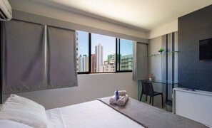 A hotel room with a bed, a desk, a chair, a TV, and a window with a city view. - Charming Studio Near the Airport Gln0805 (Recife)