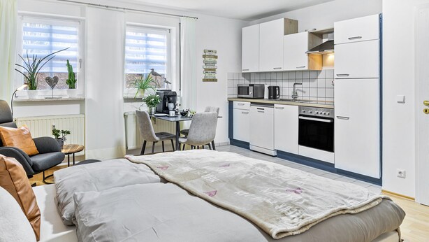 Habitación de hotel compacta con cama, un pequeño escritorio, kitchenette y ventana con persianas.