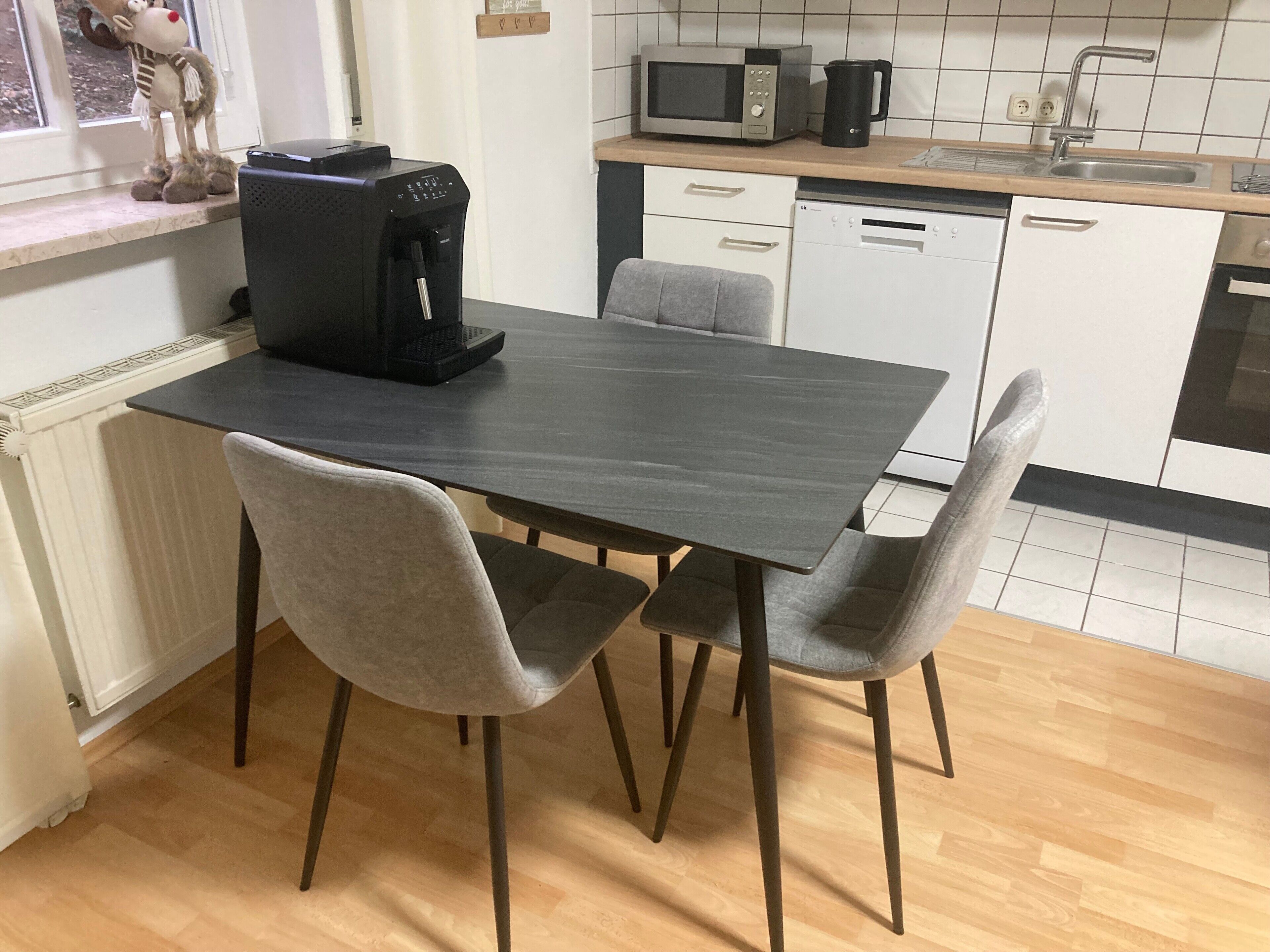 A kitchen with a dark wooden table, two grey chairs, a coffee maker, a microwave, and a dishwasher.