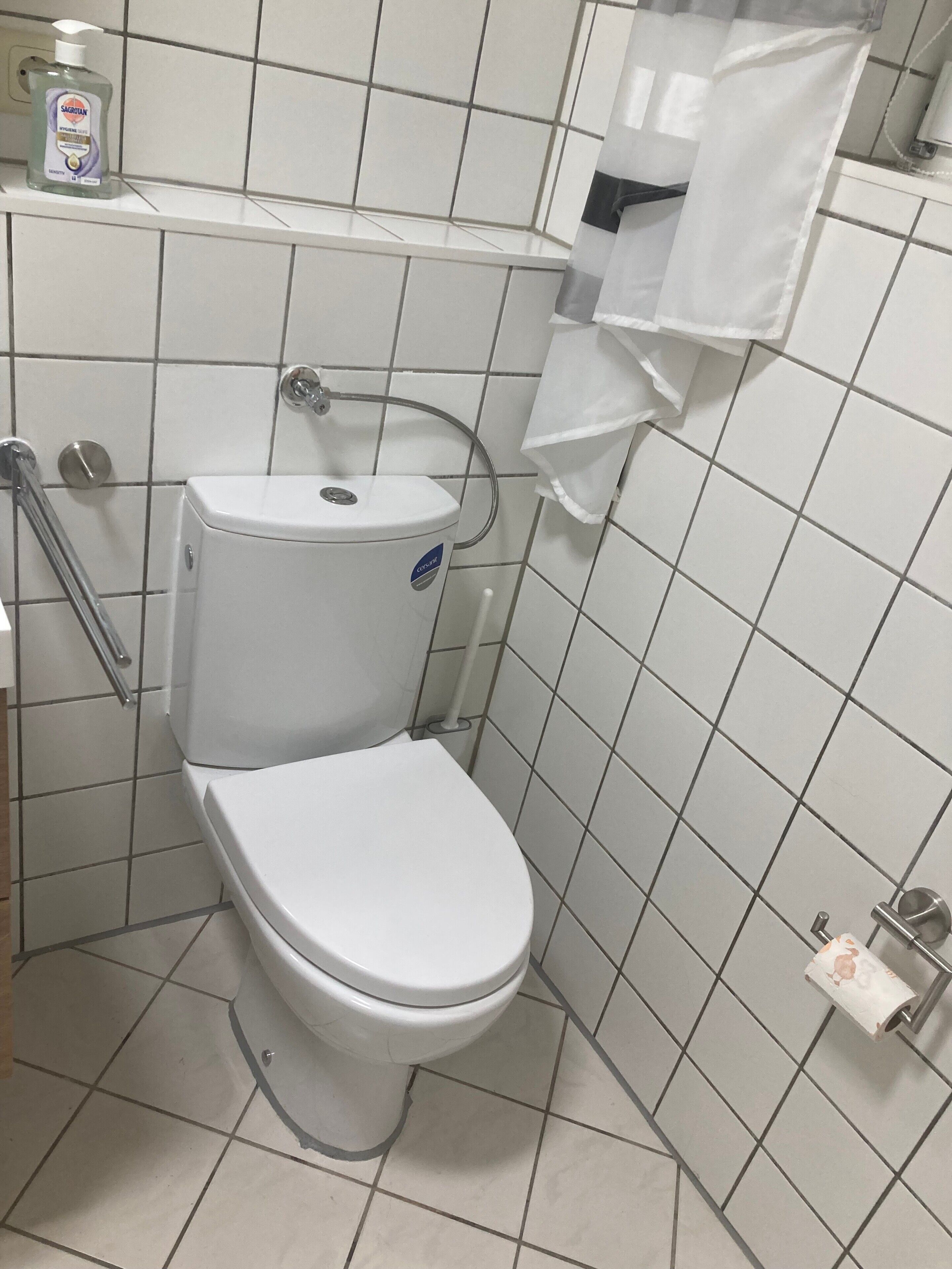 Un WC bianco con funzione bidet, maniglia di sostegno e dispenser di sapone a parete.