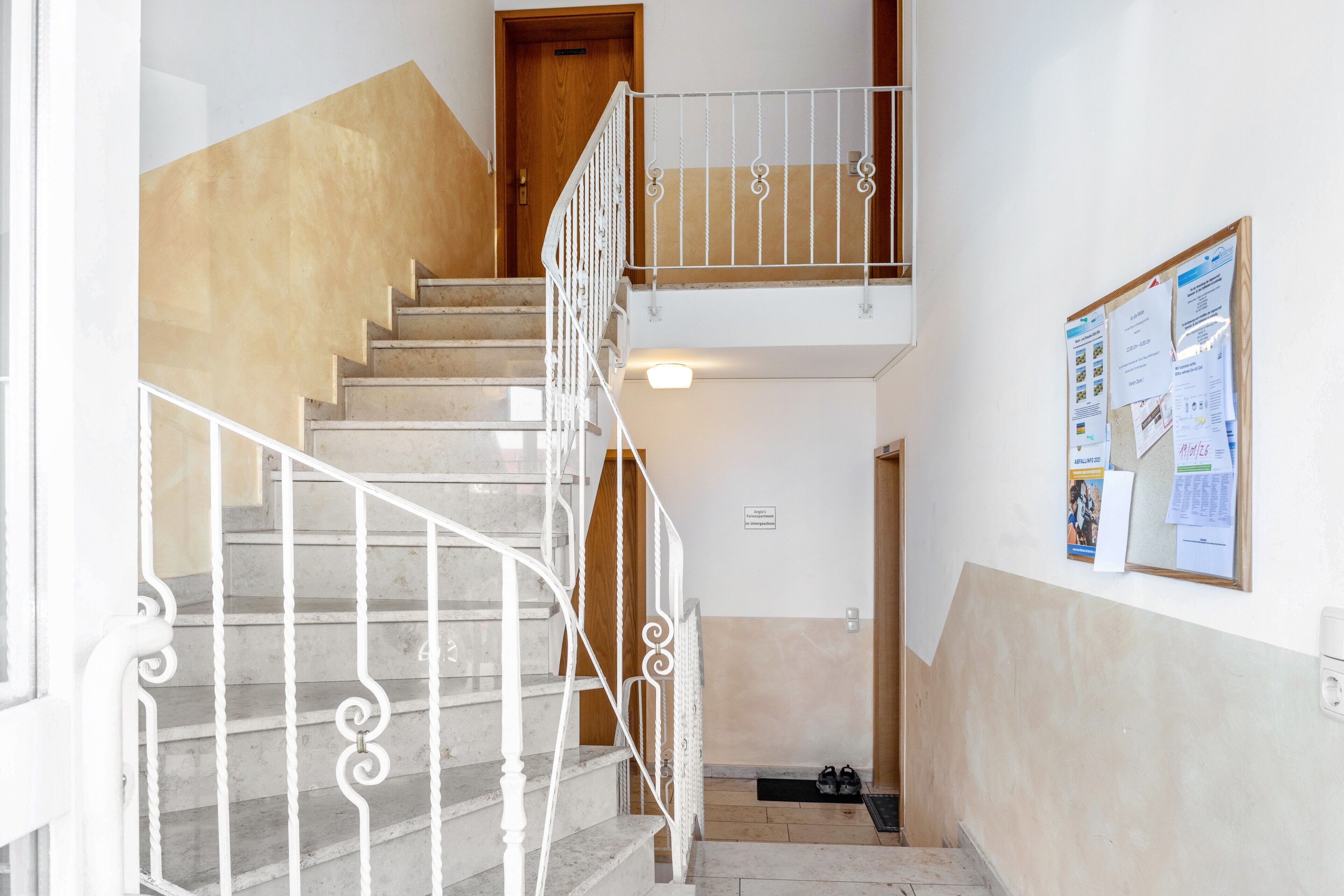 Una escalera con barandillas blancas, una puerta de madera en la parte superior y un tablón de anuncios con documentos en la pared.