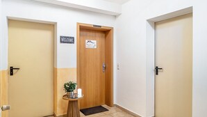 Un pasillo con tres puertas, una con la leyenda 'WELCOME' y otra con un letrero que dice 'Angie's'. Una pequeña mesa con una planta y velas se encuentra en el centro.