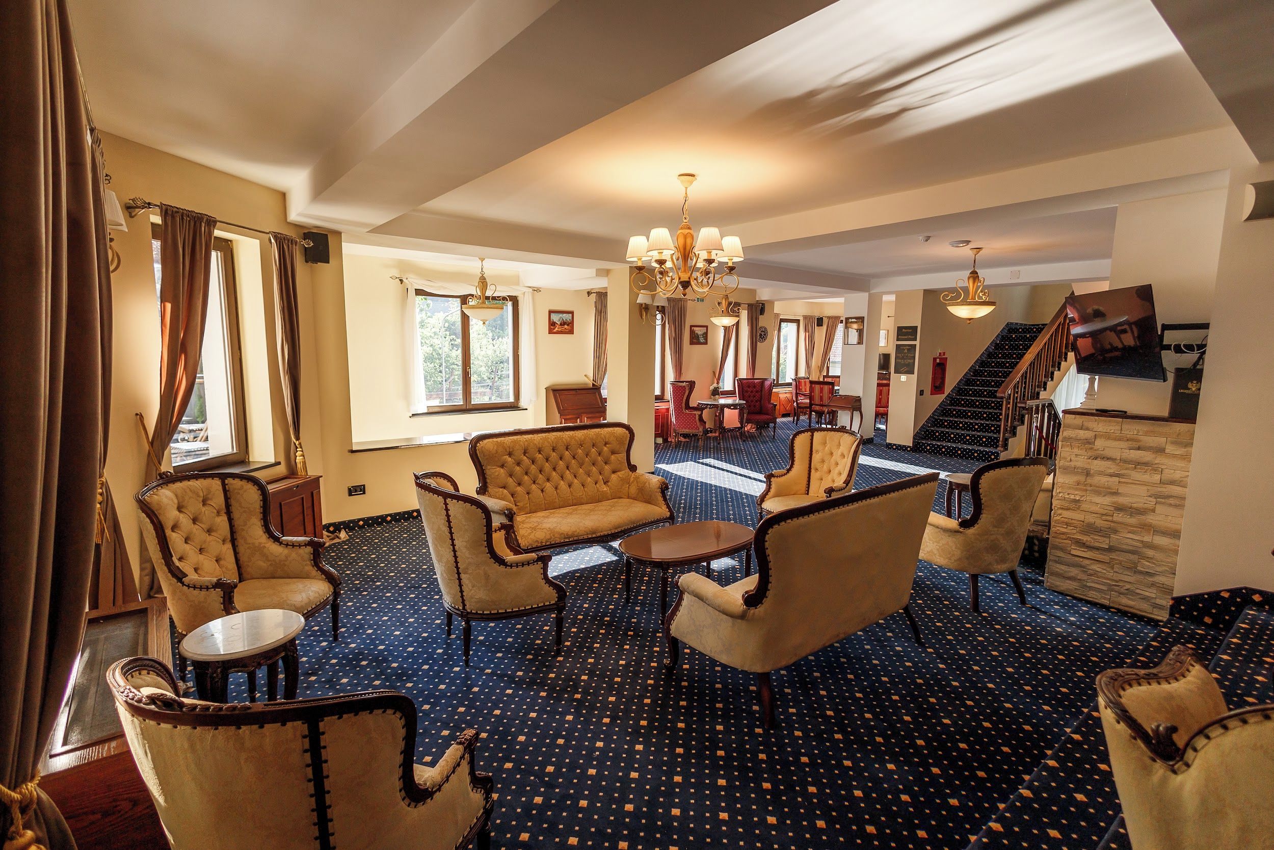 A hotel lobby with a patterned carpet, seating area with armchairs and a sofa, a chandelier, and a staircase leading to another level.
