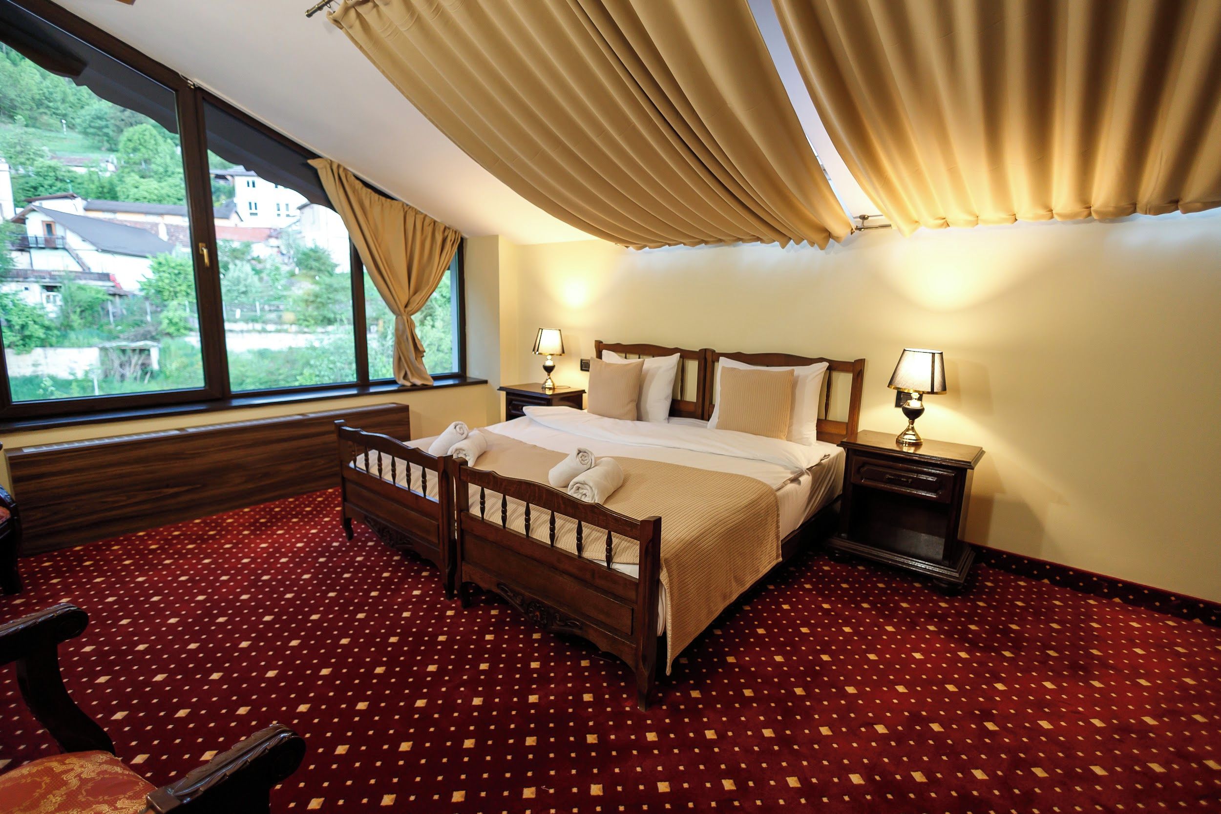 A hotel room with a large bed, two bedside tables with lamps, a wooden headboard, a red patterned carpet, and a view of houses through the window.