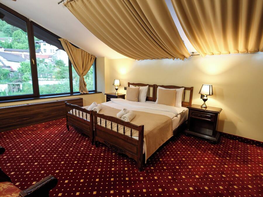 A hotel room with a large bed, two bedside tables with lamps, a wooden headboard, a red patterned carpet, and a view of houses through the window.
