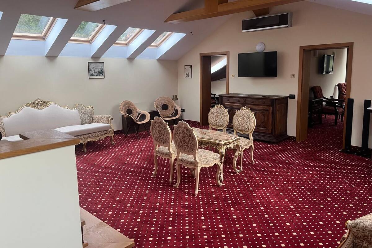 Egy piros szőnyeggel, fehér kanapéval, étkezőasztallal és székekkel, televízióval, valamint tetőablakokkal felszerelt szoba.