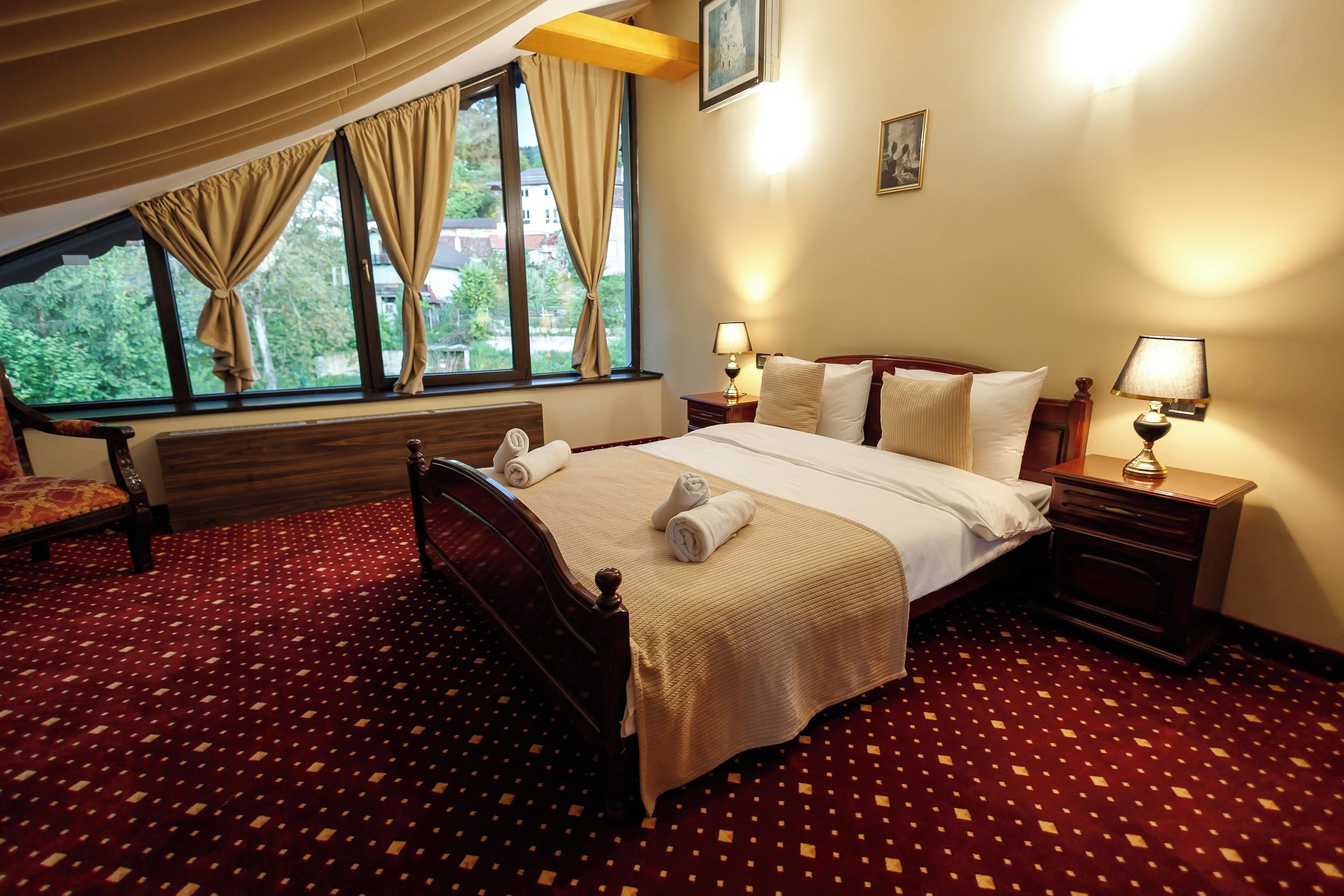 Habitación de hotel con una cama grande, dos mesitas de noche con lámparas, una ventana con cortinas y vistas a zonas verdes.