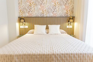 Un lit bien fait, recouvert d’une couverture blanche à texture intéressante, flanqué de deux lampes de chevet et agrémenté d’un luminaire fixé au mur.