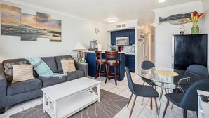 A modern living room with a sofa, coffee table, and dining area with chairs. A kitchen with blue cabinets and a microwave is visible in the background. - Ocean View Top Floor - 2 Pools - Hot Tub - GYM - Tennis (Galveston)
