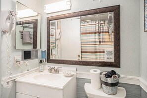 Une salle de bain avec un lavabo blanc, un grand miroir, un sèche-cheveux, un porte-serviettes et des toilettes avec un support à serviettes.