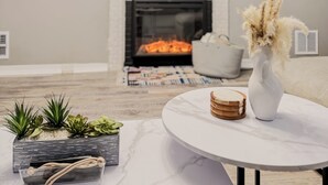 A modern living room with a fireplace, a flat-screen TV mounted above it, and a round coffee table with decorative items. - Cozy Winter Escape - Hot Tub, Game Room, Fireplace, Sleeps 6 (Waterbury)