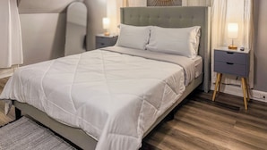 A neatly made bed with a quilted white duvet, a grey upholstered headboard, and a bedside table with a lamp. - Cozy Winter Escape - Hot Tub, Game Room, Fireplace, Sleeps 6 (Waterbury)