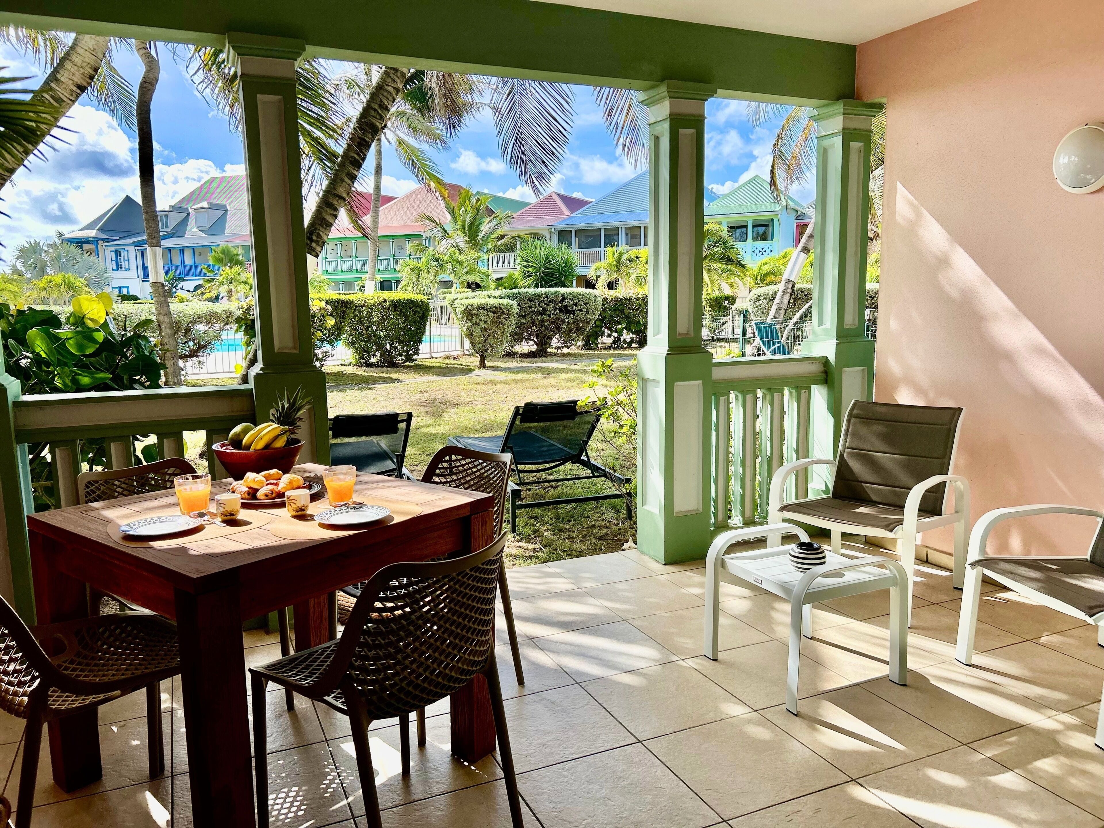 Un'area pranzo all'aperto coperta, con un tavolo apparecchiato per due, sedie e vista su una piscina e degli edifici.
