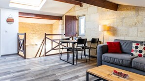 Una sala de estar moderna con pared de piedra, vigas de madera, un sofá con un cojín rojo, una mesa de comedor con sillas y un tragaluz.