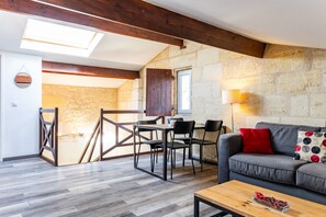 Un salon moderne avec un mur en pierre, des poutres en bois, un canapé agrémenté d’un coussin rouge, une table à manger avec des chaises et une lucarne.