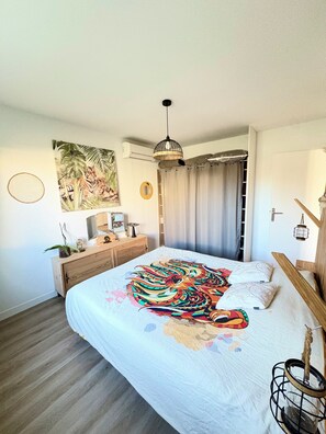 A bedroom with a bed, a wooden dresser, a wall-mounted air conditioner, a hanging light fixture, and a large painting.