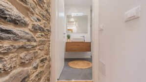 Un couloir avec un mur en pierre, un plancher en bois, un tapis gris, un lavabo blanc et un miroir.
