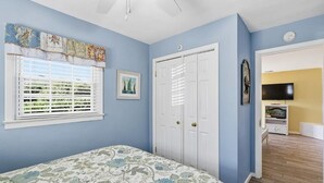 A bedroom with a bed, a window with blinds, a door, and a television in the adjacent room.