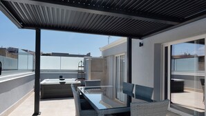 Un espace extérieur abrité, doté d’une table et de chaises, d’une paroi en verre et offrant une vue sur le paysage urbain.