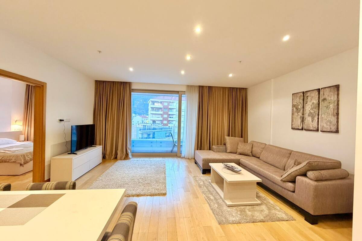 Quarto de hotel moderno com sofá, TV, mesa de jantar e varanda com vista para edifícios.