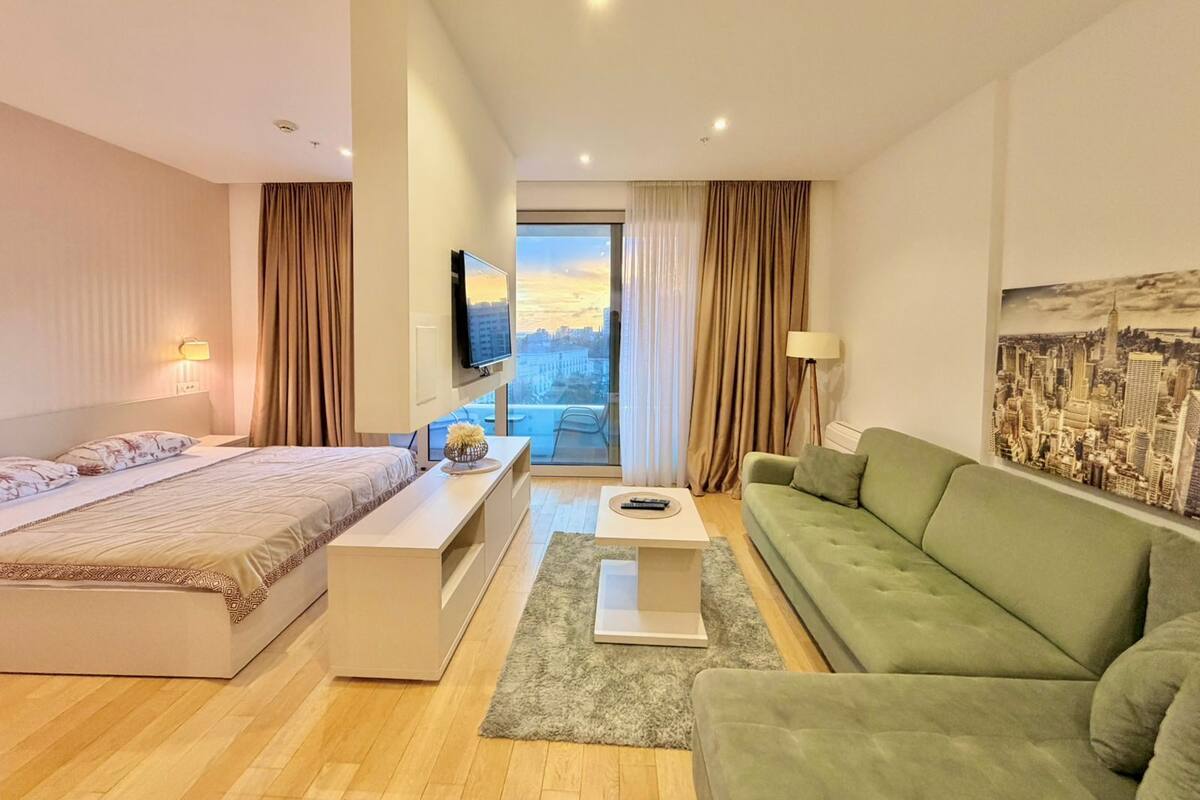 Quarto de hotel moderno com uma cama, um sofá verde, uma TV e vista para a cidade.