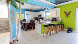 A modern kitchen with white cabinets, a green accent wall, a dining area with a wooden table and chairs, and a large island with bar stools. - Conch Nation – 4 Bedroom / 3 Bath Gulfside Home with Dockage (Marathon)