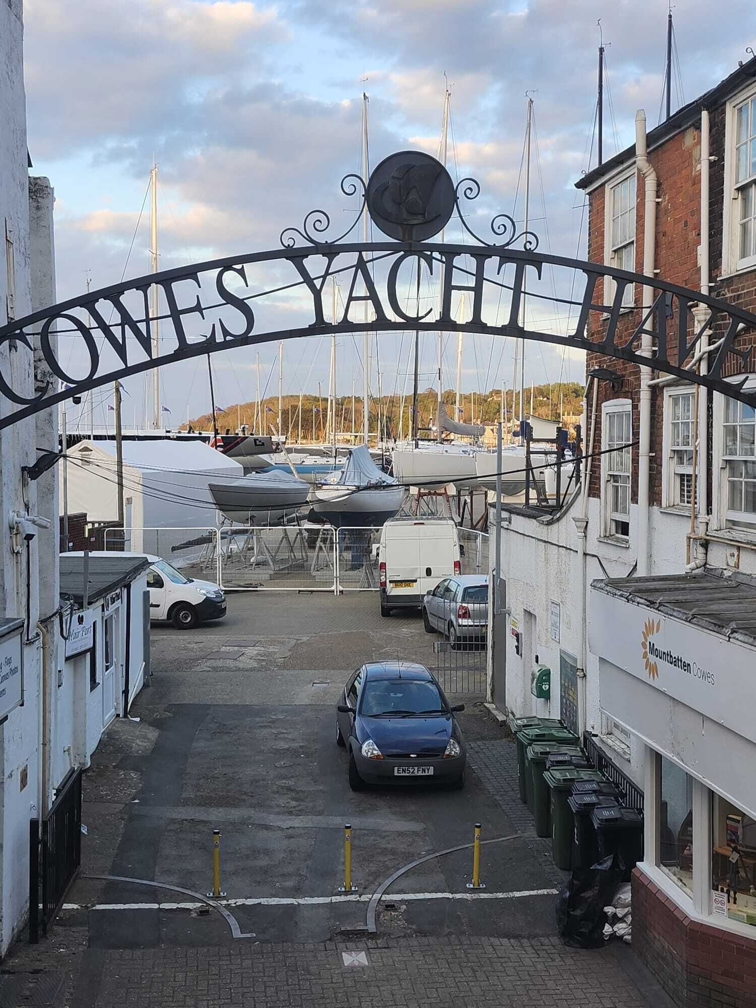 Der Eingang einer Marina mit einem Schild, auf dem 'OWES YACHTS' steht, und einem parkenden Auto im Vordergrund.
