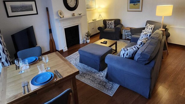 Un salon avec une cheminée, un canapé, une table à manger dressée pour deux personnes et une horloge au mur.