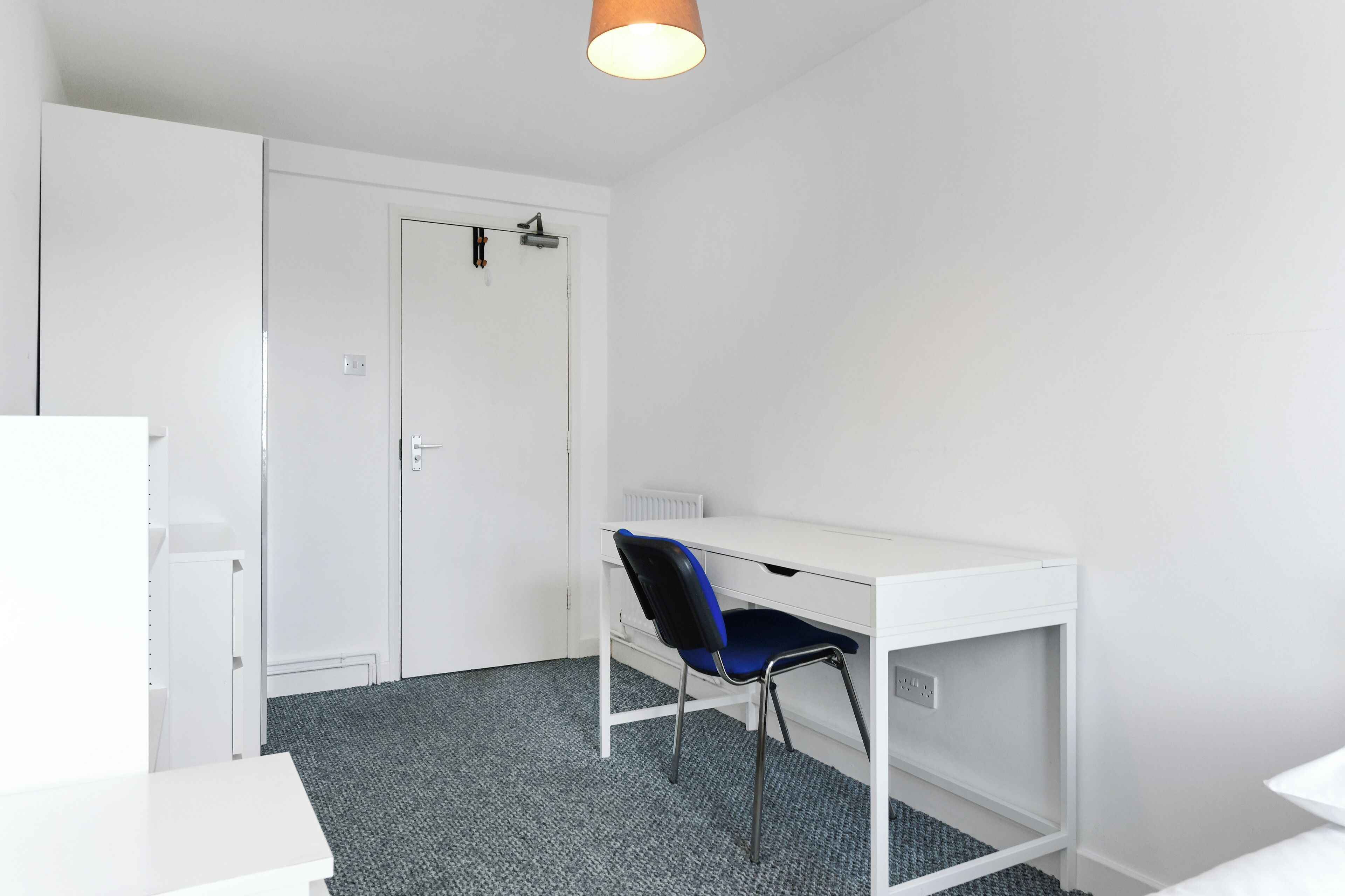 Una habitación pequeña y blanca con un escritorio, una silla, un radiador y una puerta.