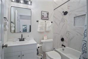A modern bathroom with a white sink, toilet, and bathtub. There are towels hanging on a rack, a framed picture on the wall, and a shower with a glass door. - Pristine 3 BR/ 2BA Abode Close to Lovely Downtown! (San Antonio)