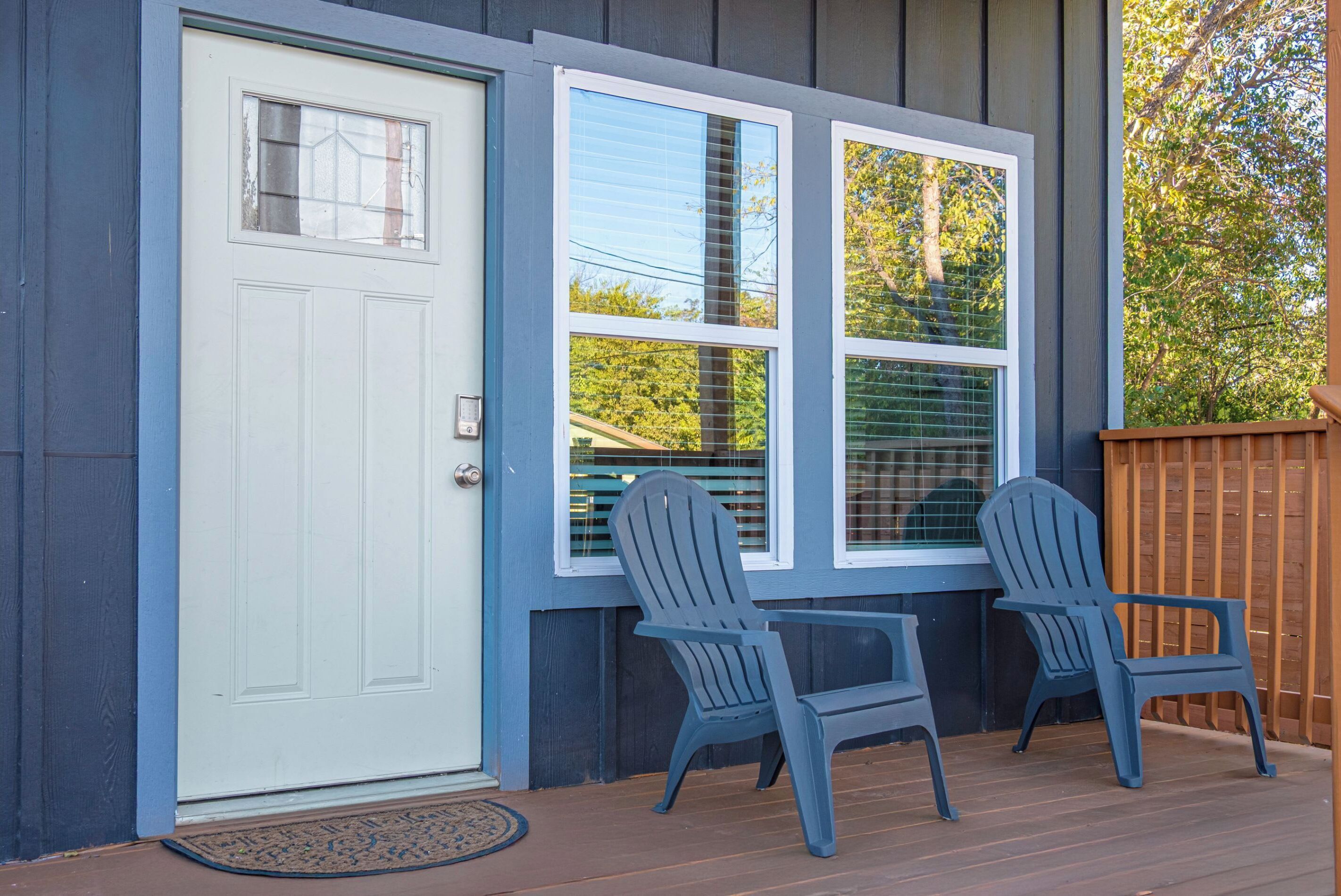 Un porche à l’avant de la maison, avec une porte blanche, deux fauteuils Adirondack et une terrasse en bois.
