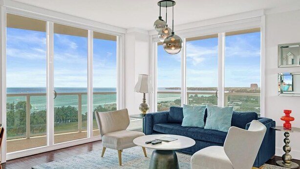 Ein geräumiges Wohnzimmer mit großem Fenster zum Meer, ein blaues Sofa, zwei Sessel, ein runder Couchtisch und eine hängende Leuchte.