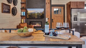 L’intérieur d’un chalet accueillant, avec une cheminée, une table à manger en bois agrémentée d’une bouteille de vin et de verres, et une cuisine moderne dotée d’un réfrigérateur en acier inoxydable.