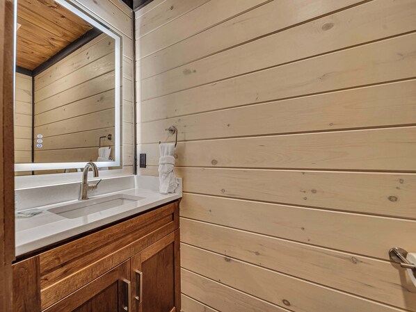 A bathroom with a wooden wall, a large mirror, a sink, and a towel rack. - Beautiful New 2 Level Build I Private Hot Tub I Fire Pit I Game Room (Sevierville)