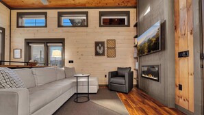 A modern living room with a wooden ceiling, a fireplace, a sofa, a chair, and a TV mounted on the wall. - Fab New 2 Level Build I Fire Pit I Private Hot Tub I Game Room (Sevierville)