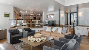 Ein geräumiges Wohnzimmer mit einer großen Couch, Sesseln und einem Couchtisch. Im Hintergrund befindet sich eine Küche mit Essbereich.