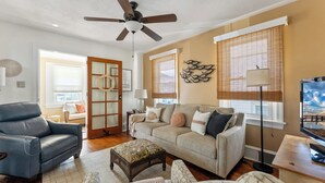 Una sala de estar con sofá, sillón y reposapiés, ventilador de techo y televisor.