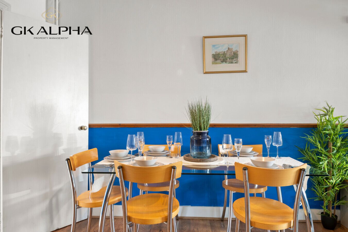 Ein Essbereich mit einem Tisch, gedeckt für sechs Personen, Stühlen, einer Vase mit Grünpflanzen und einem gerahmten Bild an der Wand.