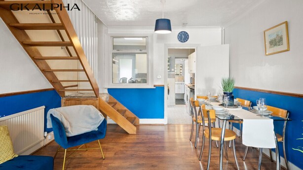A modern kitchen with a blue accent wall, wooden staircase, and a dining area with a table set for four. - Heartwarming Windsor Cottage with Free Parking (Berkshire)