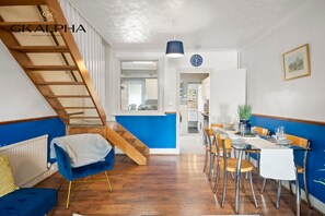 A modern kitchen with a blue accent wall, wooden staircase, and a dining area with a table set for four. - Heartwarming Windsor Cottage with Free Parking (Berkshire)
