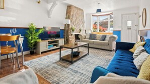 A living room with a blue sofa, a coffee table, a TV on a stand, a stone wall, and a window with a view of a garden. - Heartwarming Windsor Cottage with Free Parking (Berkshire)