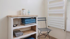 Une pièce avec un radiateur blanc, une étagère où sont pliées des serviettes, une petite table avec une bouteille et une chaise.