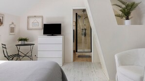 Une chambre à coucher comprenant un lit, une télévision, une petite table avec une plante, une chaise, une commode blanche et un espace douche.