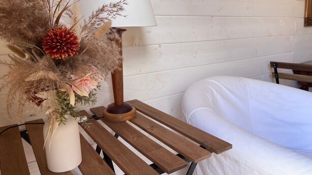 Une table de chevet en bois sur laquelle se trouve un vase blanc contenant des fleurs séchées et une lampe.